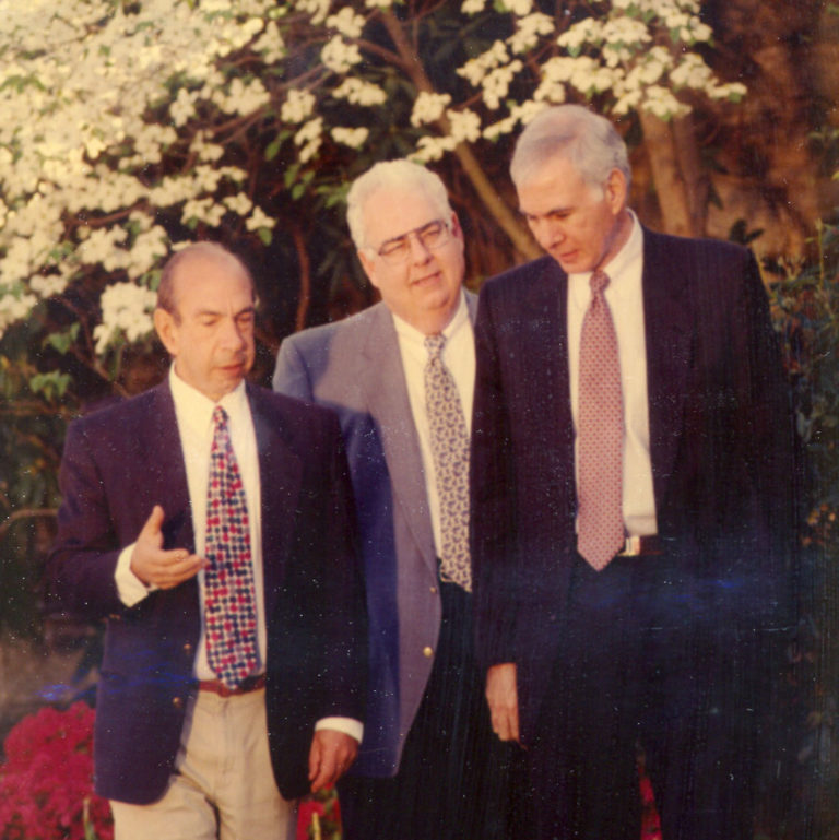 Three men in suits, the founders of COSEHQ and a board member, walking and talking outside with blooming tree in the background.